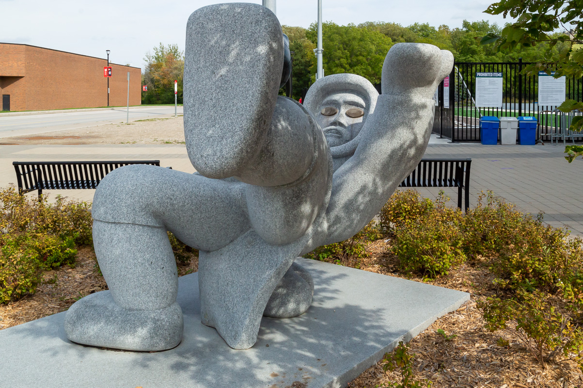 A stone sculpture of a young Inuk soccer player performing an Alaskan high kick, an official Inuit sport of the Arctic Winter Games.